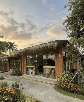 Tropical Bali furniture showroom exterior with thatched roof, floor-to-ceiling glass display windows, and surrounding flower garden.